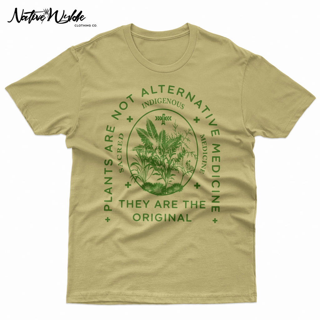 A natural sand-colored T-shirt with a dark green graphic print on the front. The graphic features a central botanical illustration inside an oval, surrounded by the text 'PLANTS ARE NOT ALTERNATIVE MEDICINE', 'INDIGENOUS', and 'THEY ARE THE ORIGINAL'.