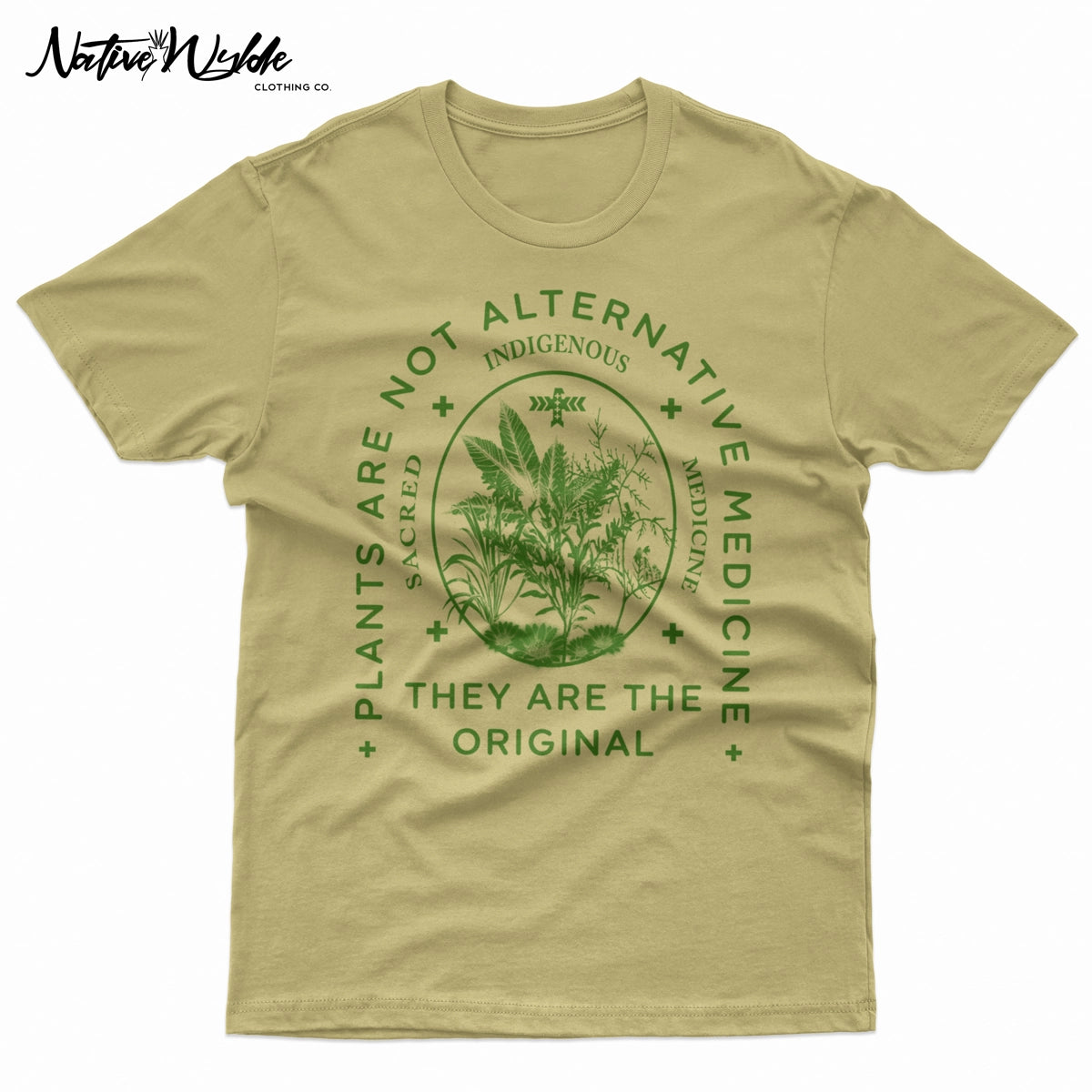 A natural sand-colored T-shirt with a dark green graphic print on the front. The graphic features a central botanical illustration inside an oval, surrounded by the text 'PLANTS ARE NOT ALTERNATIVE MEDICINE', 'INDIGENOUS', and 'THEY ARE THE ORIGINAL'.