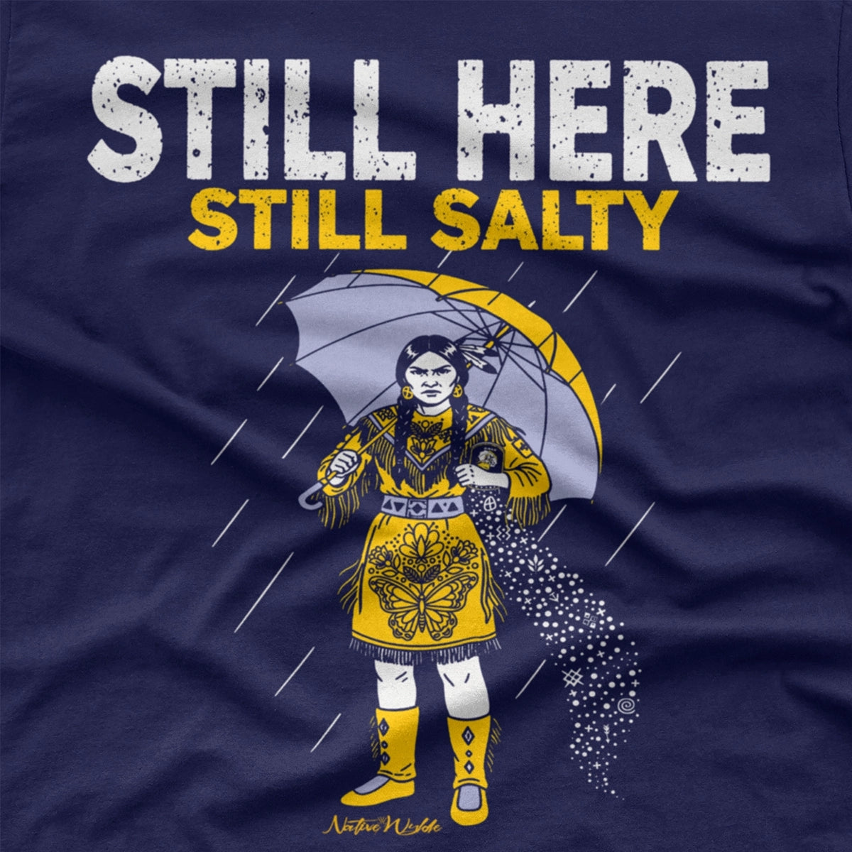 A navy blue short-sleeve T-shirt with a graphic print of a woman in a yellow dress holding an umbrella in the rain, with the text 'STILL HERE STILL SALTY' in white and yellow. 