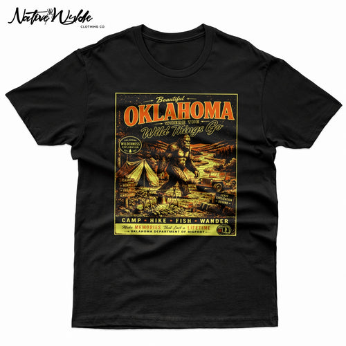 A vintage-style Oklahoma travel poster graphic printed on a black shirt. The design features large orange-red 'OKLAHOMA' lettering, a Bigfoot scene with a tent, jeep, river, and campfire, plus yellow text reading 'Where the Wild Things Go' and 'Make Memories That Last a Lifetime'.