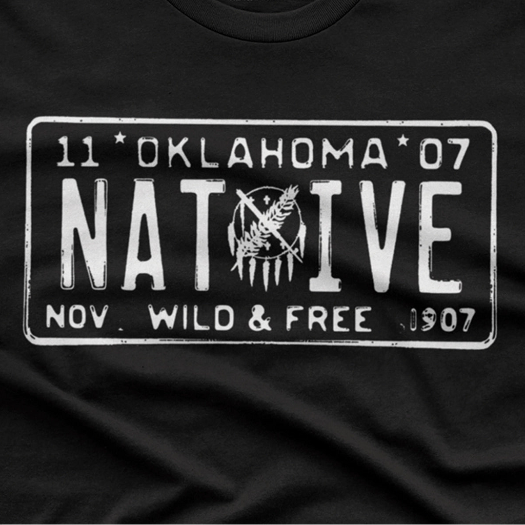 Close-up of a black T-shirt with a large distressed white graphic styled like an Oklahoma license plate reading "NATIVE" with "11 Oklahoma 07" and "Nov Wild & Free 1907".