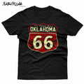 A close-up of a black shirt featuring a large graphic print of the Oklahoma Route 66 shield. The design has bonecolored text and numbers outlined in red, with 'The Mother Road OKLAHOMA' at the top and 'Oklahoma City Tulsa' in the background shield, along with other Oklahoma cities Route 66 travel through.