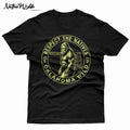 A black short-sleeve crewneck t-shirt featuring a circular graphic print of Bigfoot in the center with the text 'RESPECT THE NATIVES' and 'OKLAHOMA WILD' in bone color around the border. 