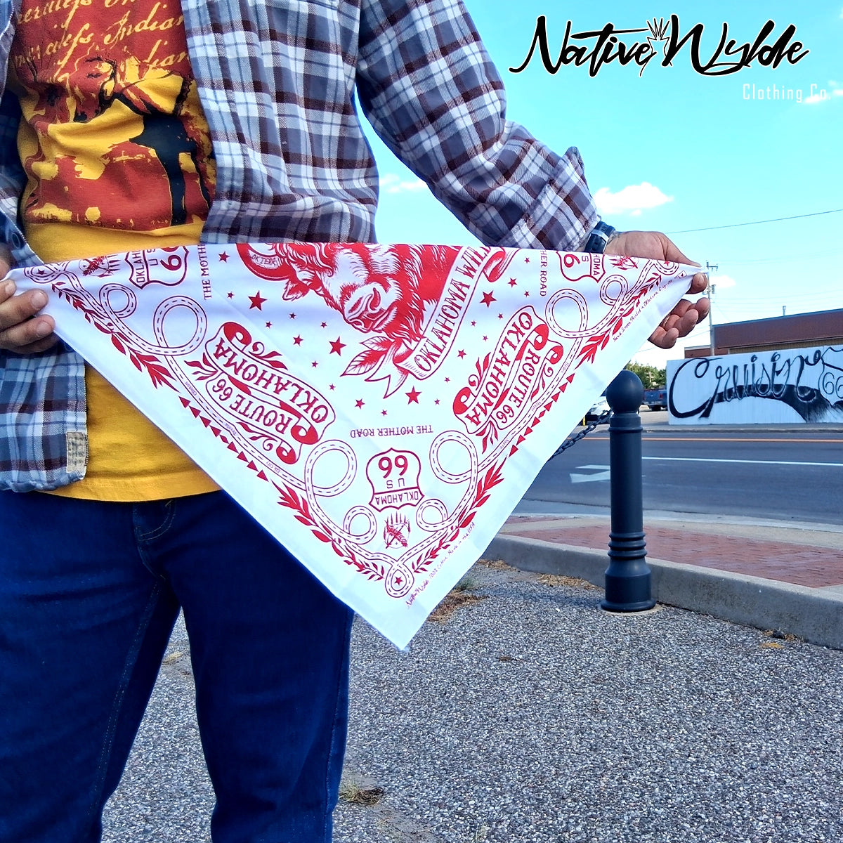 A person outdoors holding up a white bandana with a red graphic print featuring Native American artwork and the text 'OKLAHOMA' and 'Route 66'. .