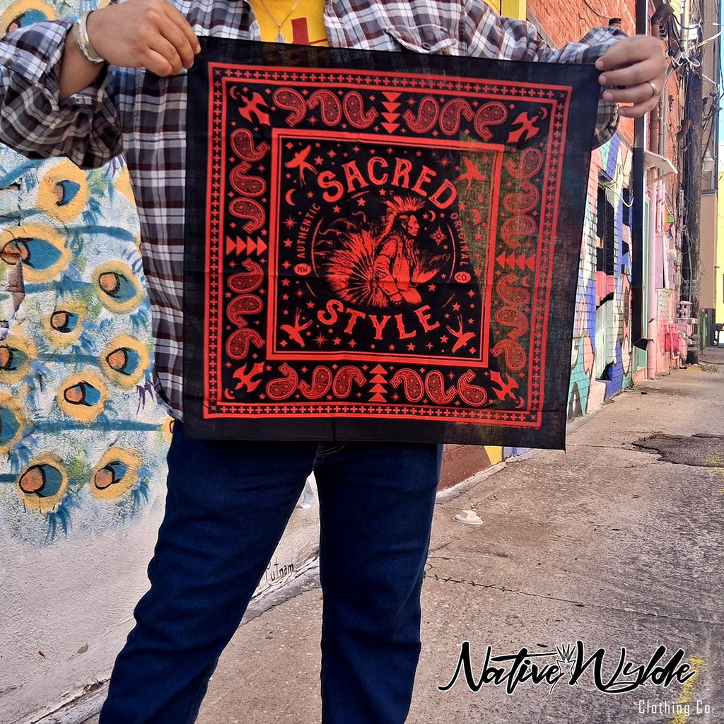 A person outdoors holding up a square black bandana-style print featuring red ornamental border details and the words “SACRED STYLE” with a central bird illustration. The person wears a plaid shirt and jeans, with a colorful mural and street buildings in the background.