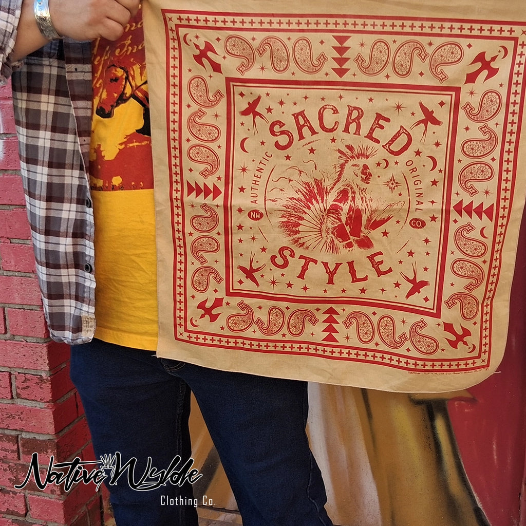 A person holding up a square tan bandana-style scarf with red western-style printed graphics and the words 'SACRED STYLE' in the center. The person is wearing a plaid shirt, yellow graphic tee, and blue jeans, standing next to a red brick wall.