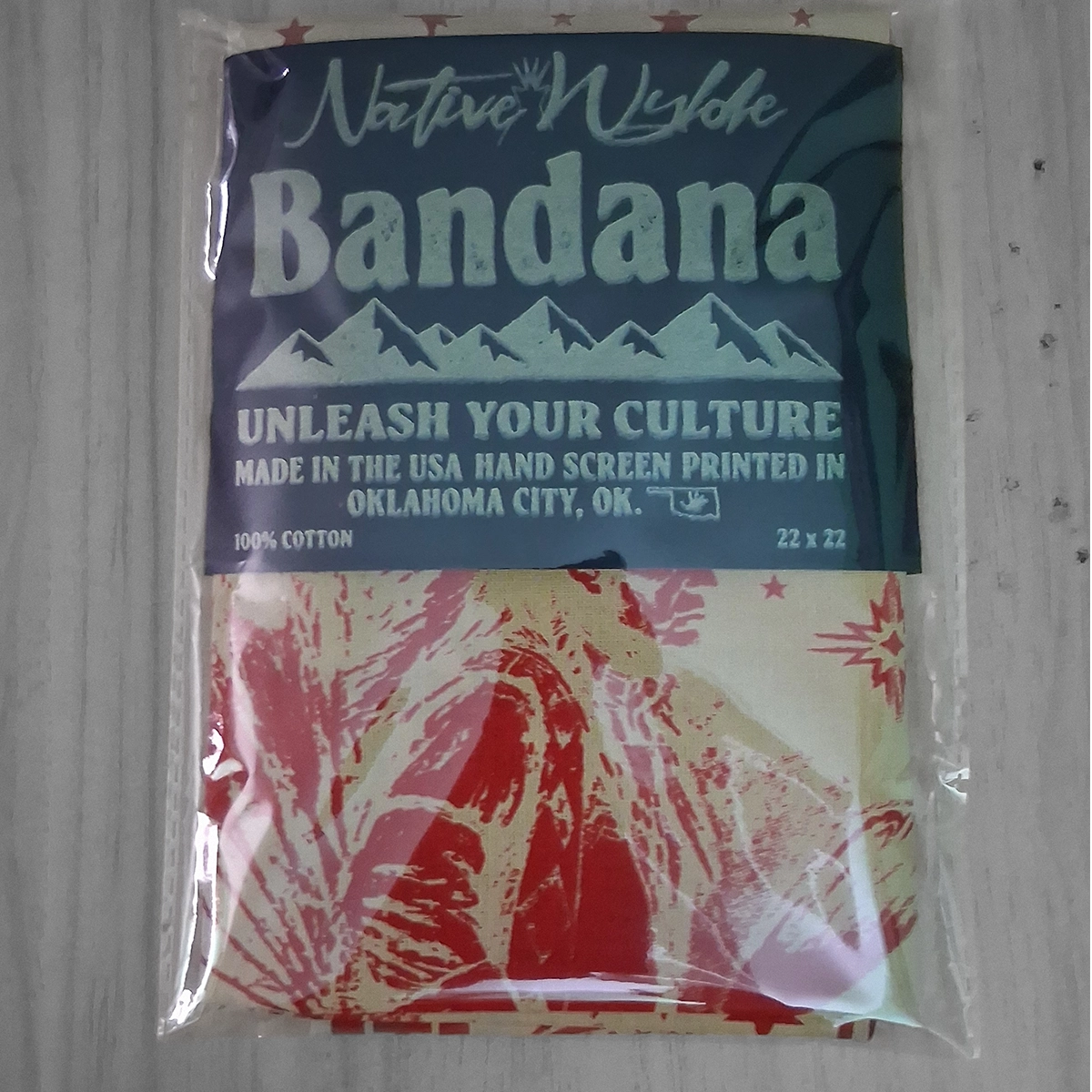 A packaged Native Wyldc Bandana featuring a  header card with white text and a red-and-cream patterned cotton bandana visible inside a clear plastic sleeve, placed on a light wood surface.