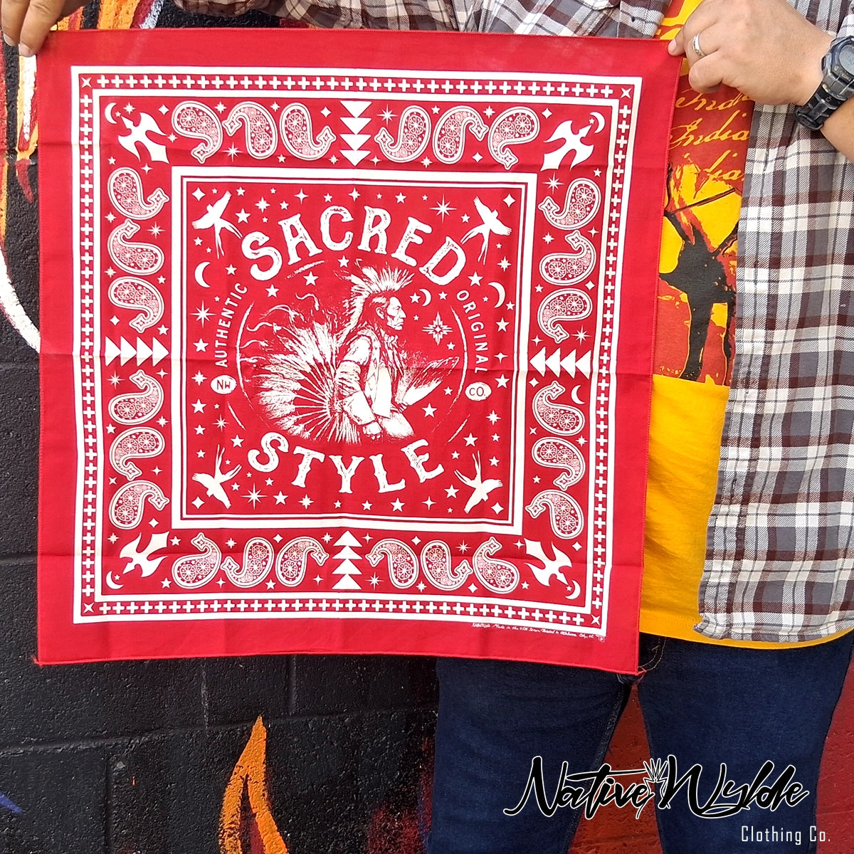 A person holds up a red bandana-style square cloth with white printed graphics and text reading “SACRED STYLE.” The design includes stars, moons, geometric borders, and a central Native American dancer illustration.