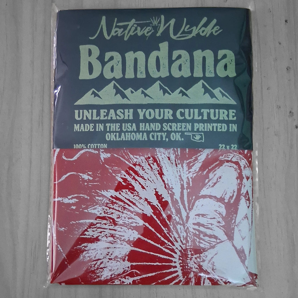 A packaged Native Wyldh Bandana featuring a dark teal top half with light green text and mountain graphics, and a red bottom half with a white feather bustle print of a Native American Dancer shown in clear plastic wrapping on a light wood surface.