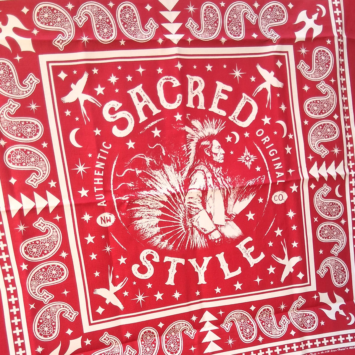 A red bandana featuring a white graphic print with the words 'SACRED STYLE' and a central Native American profile illustration, surrounded by stars, moons, and geometric border patterns.