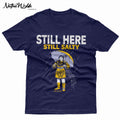 A navy blue short-sleeve T-shirt with a graphic print of a woman in a yellow dress holding an umbrella in the rain, with the text 'STILL HERE STILL SALTY' in white and yellow. 