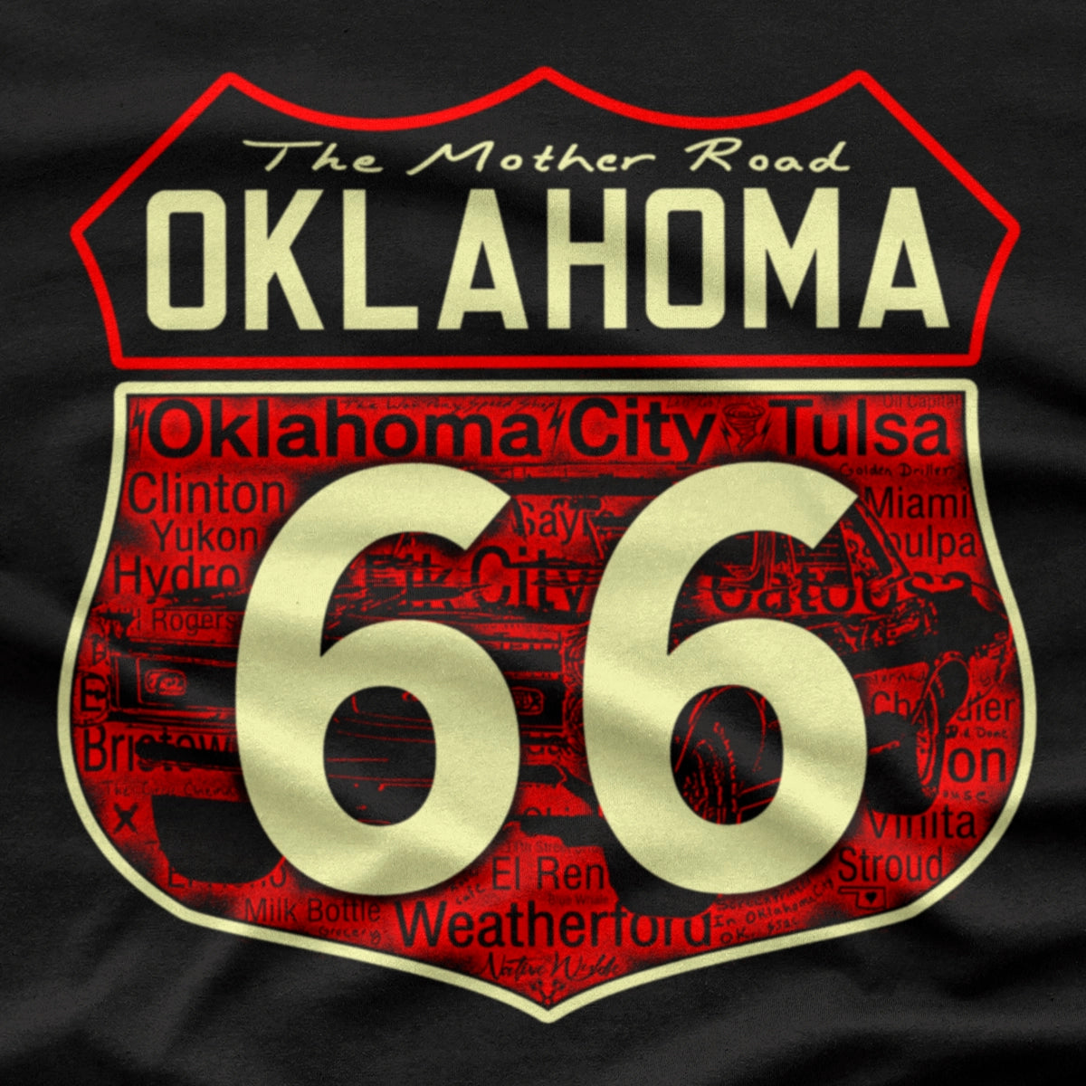 A close-up of a black shirt featuring a large graphic print of the Oklahoma Route 66 shield. The design has bonecolored text and numbers outlined in red, with 'The Mother Road OKLAHOMA' at the top and 'Oklahoma City Tulsa' in the background shield, along with other Oklahoma cities Route 66 travel through.