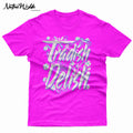 A bright pink crewneck t-shirt featuring a large graphic on the front that reads 'Tradish Delish' in stylized white and light blue lettering with small floral accents and a Native American Shawl Dancer.